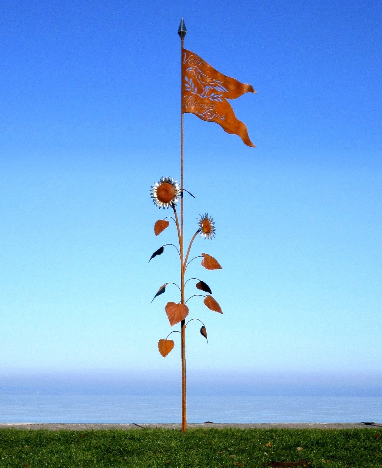 Künstlerische Metall-Stele am See: Eine stilisierte Sonnenblume aus Metall mit einer orangefarbenen Fahne an der Spitze vor blauem Hintergrund.