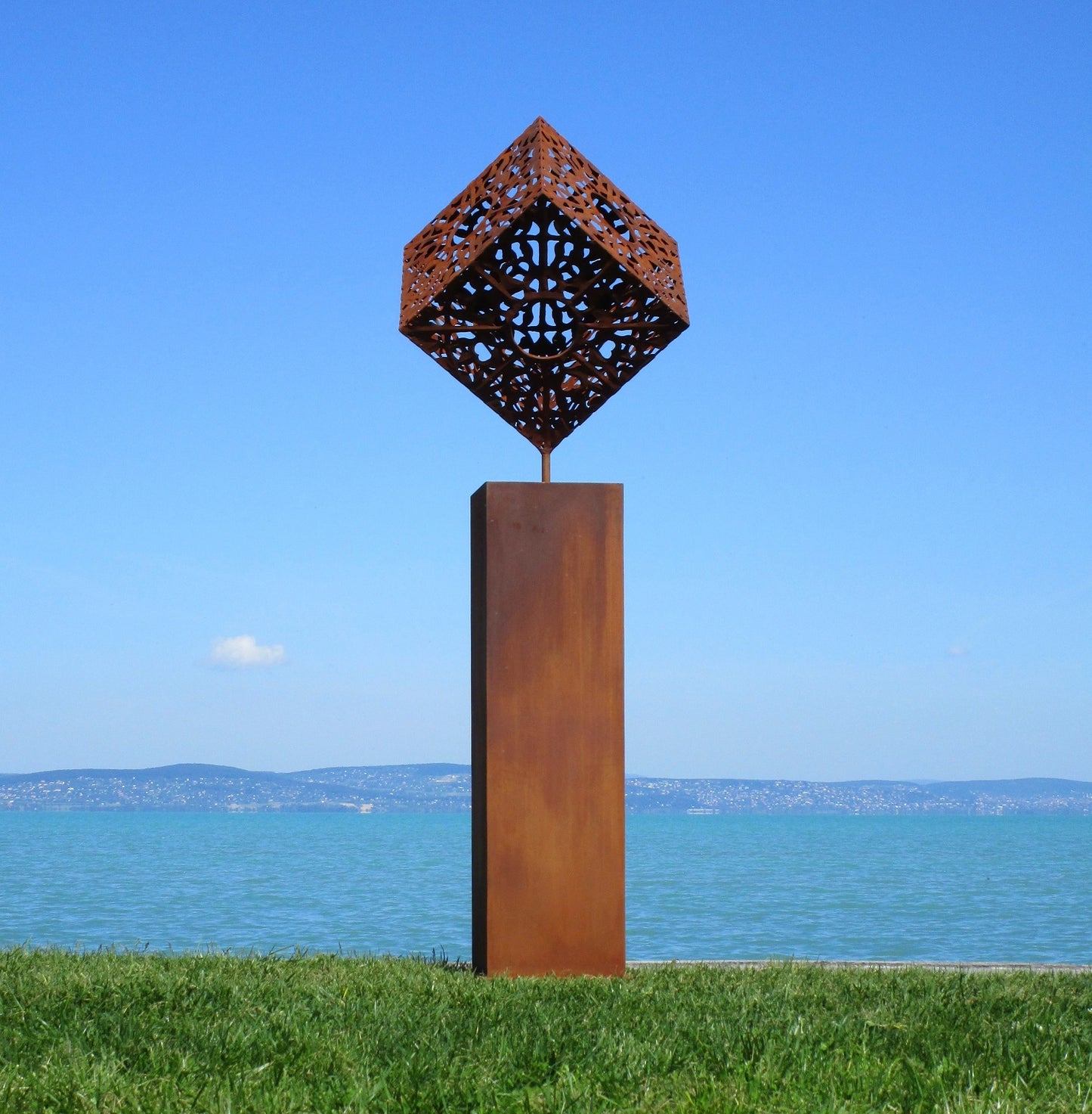 Filigrane Würfel-Skulptur aus Metall auf einer hohen, rostfarbenen Säule vor einem weiten Panorama-Blick auf einen blauen See.