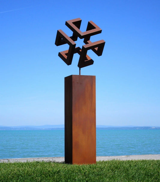 Abstrakte geometrische Skulptur aus Cortenstahl: Ein verschlungenes, würfelähnliches Symbol in markanter Rost-Optik, montiert auf einer hohen rechteckigen Stele direkt am Seeufer unter strahlend blauem Himmel.