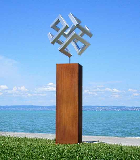 Abstrakte geometrische Skulptur aus Cortenstahl: Ein verschlungenes, würfelähnliches Symbol in markanter Rost-Optik, montiert auf einer hohen rechteckigen Stele direkt am Seeufer unter strahlend blauem Himmel.
