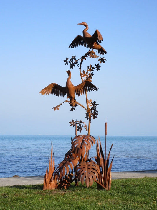 Zwei Kormoran-Skulpturen aus Cortenstahl: Eine vertikale Metall-Installation mit zwei Vögeln in charakteristischer Pose auf einem verzweigten Ast, thronend über einer Basis aus filigranem Schilf und Monstera-Blättern in markanter Rost-Optik am Seeufer.