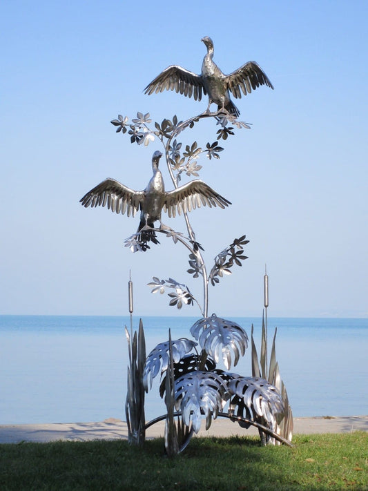 Zwei Kormoran-Skulpturen aus glänzendem Edelstahl: Eine vertikale Installation mit zwei Metallvögeln in Pose auf einem verzweigten Ast, thronend über einer Basis aus filigranem Schilf und Monstera-Blättern direkt am Seeufer.