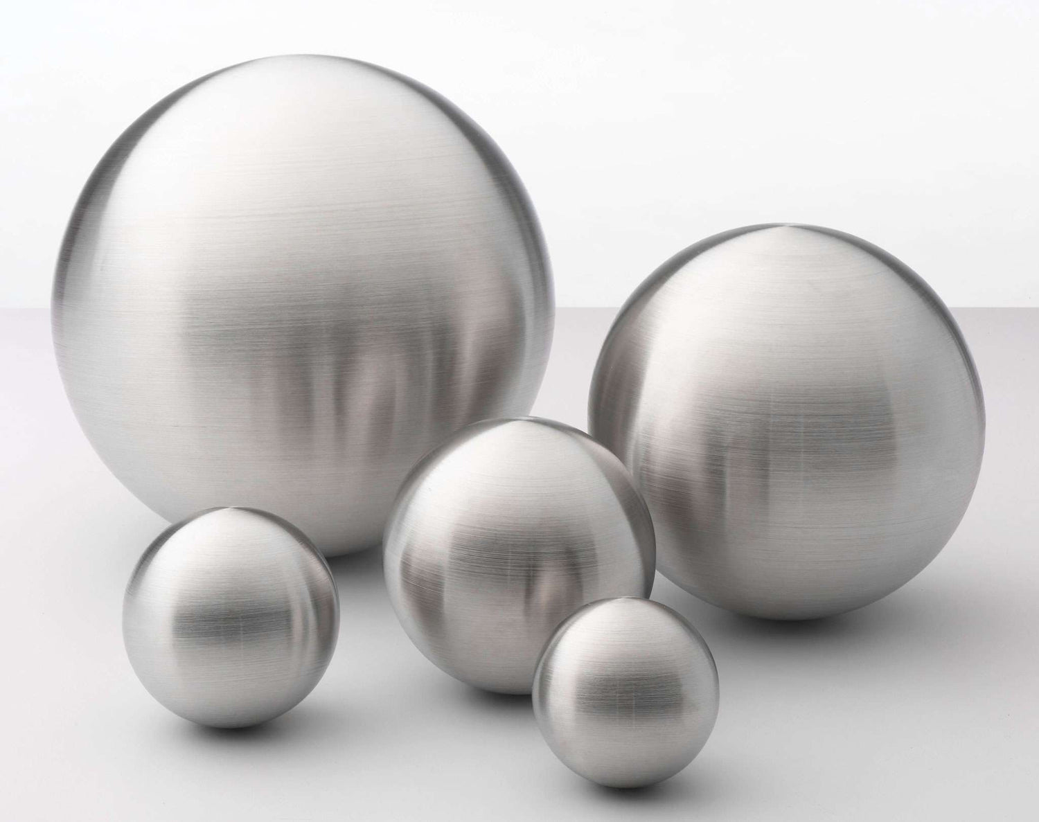 Dieses Bild präsentiert eine Gruppe von fünf perfekt geformten Kugeln aus gebürstetem Metall, die auf einer neutralen, hellen Oberfläche arrangiert sind. Die Kugeln haben unterschiedliche Größen – von einer sehr großen, dominanten Kugel im Hintergrund bis hin zu kleinen Exemplaren im Vordergrund – und wirken durch ihre matte, silbrig-graue Textur sehr edel. Das Licht fällt weich von der Seite ein, was die Rundungen der Kugeln betont und feine horizontale Schleifspuren auf dem Metall sichtbar macht. Durch di