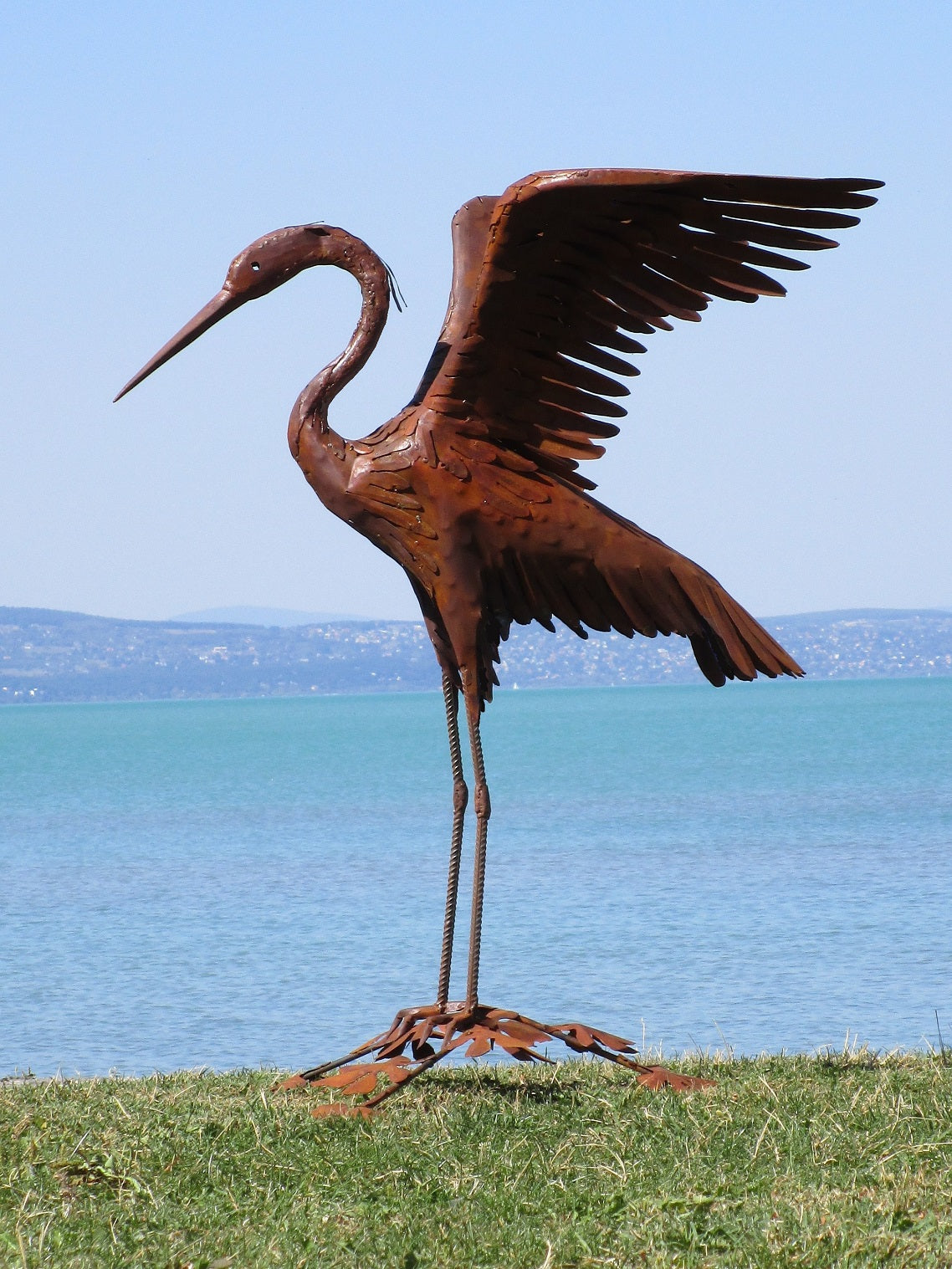 Elegante Reiher-Skulptur aus Cortenstahl: Ein filigraner Metall-Vogel mit ausgebreiteten Flügeln und langem Schnabel in Rost-Optik am Seeufer.