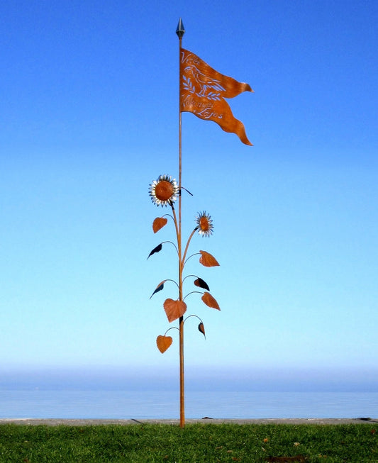Künstlerische Metall-Stele am See: Eine stilisierte Sonnenblume aus Metall mit einer orangefarbenen Fahne an der Spitze vor blauem Hintergrund.
