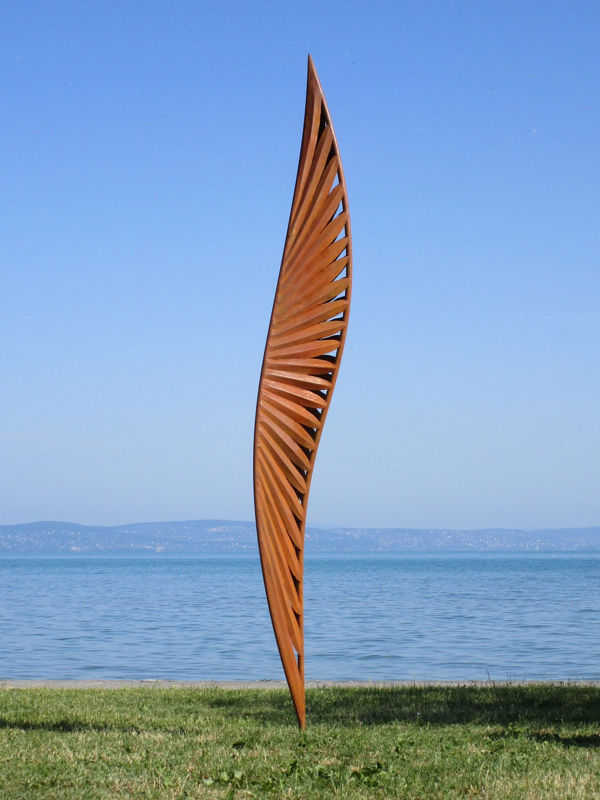 Großformatige Skulptur aus Cortenstahl: Ein quadratisches Kunstwerk mit floralen Durchbrüchen vor einem See-Panorama und fernen Bergen.