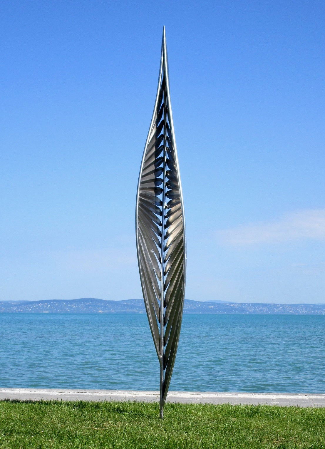 Detailansicht einer skulpturalen Metall-Installation: Ein auf der Spitze stehender Würfel mit komplexen Cut-Out-Mustern auf einer grünen Wiese vor blauem Wasser.