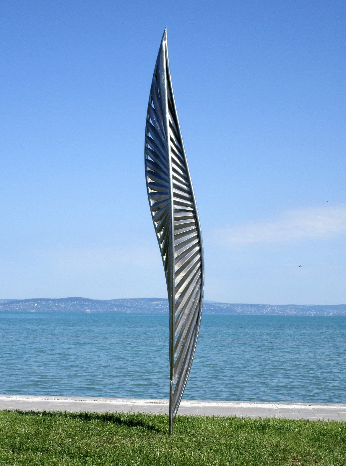 Detailansicht einer skulpturalen Metall-Installation: Ein auf der Spitze stehender Würfel mit komplexen Cut-Out-Mustern auf einer grünen Wiese vor blauem Wasser.