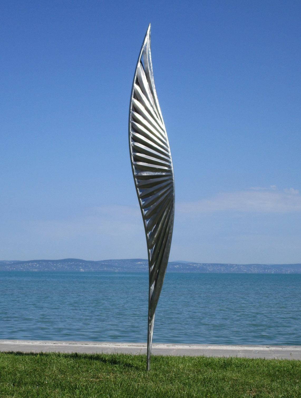 Moderne Edelstahl-Skulptur: Ein silberner, filigran durchbrochener Metallwürfel auf einer hohen Cortenstahl-Stele am Seeufer unter blauem Himmel.