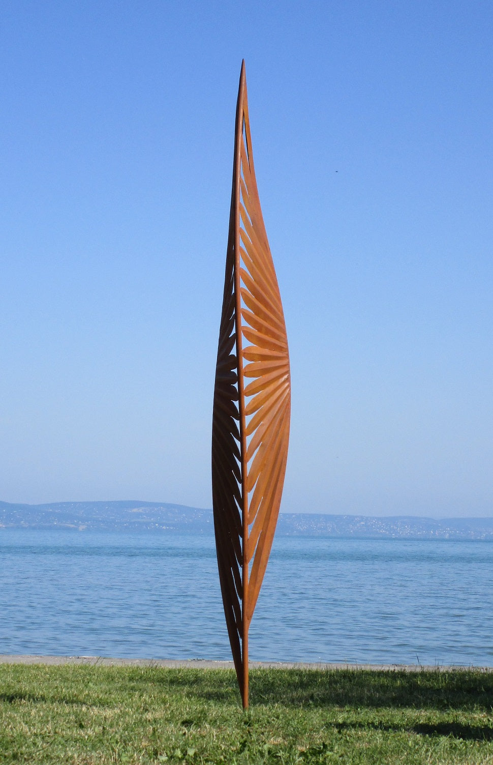 Detailansicht einer skulpturalen Metall-Installation: Ein auf der Spitze stehender Würfel mit komplexen Cut-Out-Mustern auf einer grünen Wiese vor blauem Wasser.