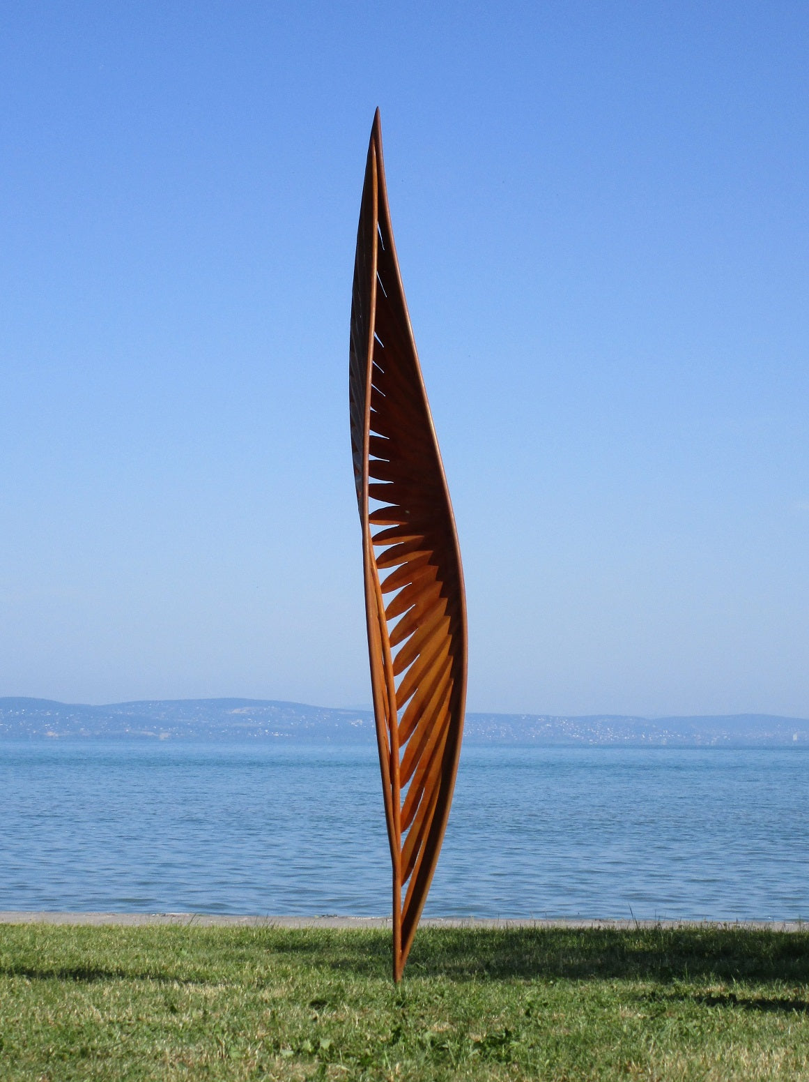 Großformatige Skulptur aus Cortenstahl: Ein quadratisches Kunstwerk mit floralen Durchbrüchen vor einem See-Panorama und fernen Bergen.