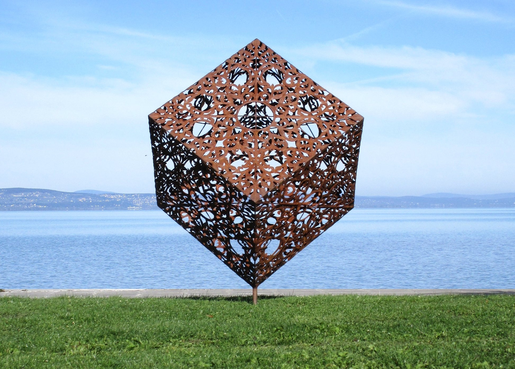 Filigraner Metallwürfel als Kunstobjekt: Eine auf der Ecke stehende Skulptur mit kunstvollen Durchbrüchen vor einem blauen Panorama-Hintergrund am See.