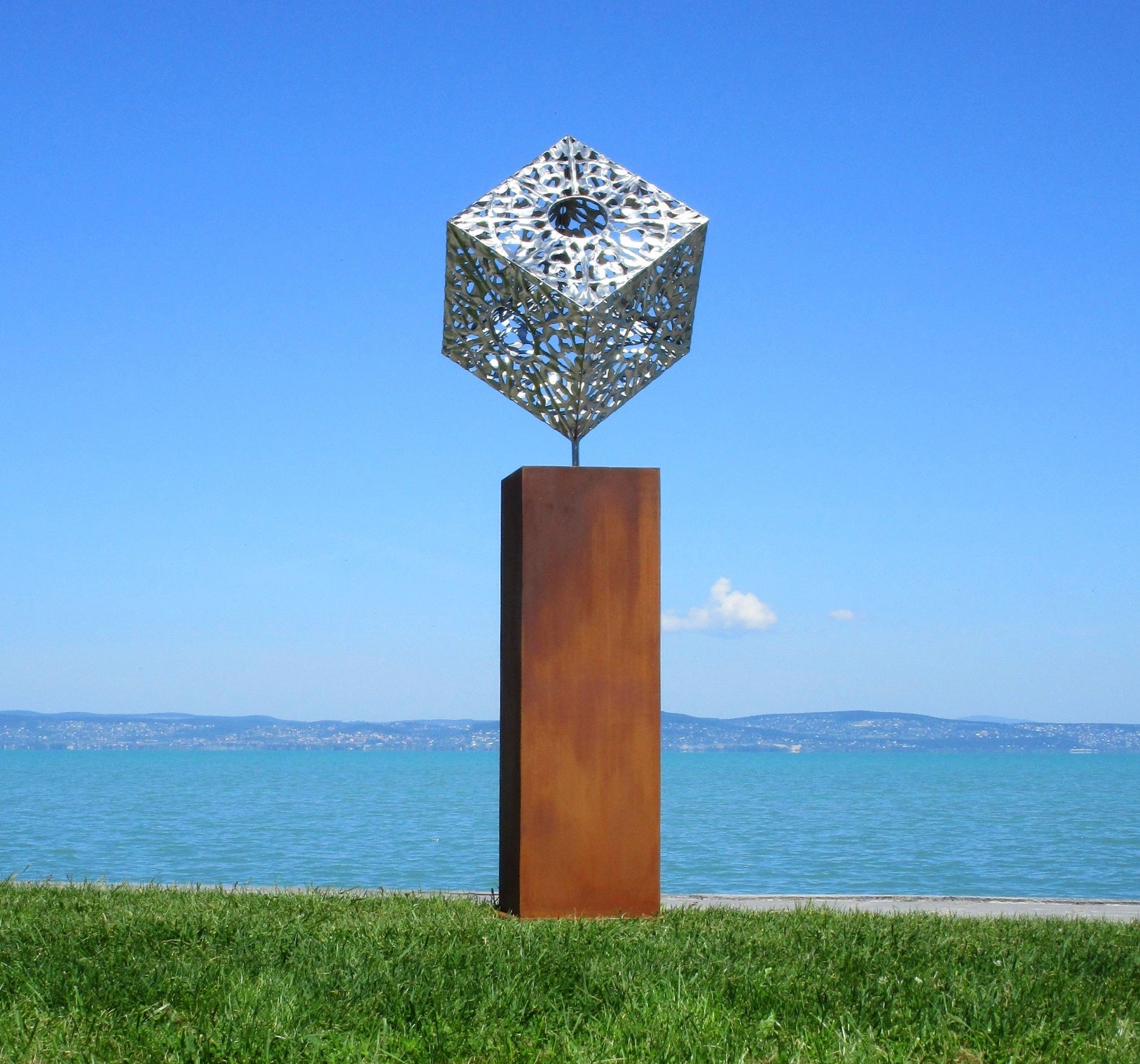 Moderne Metallskulptur in Würfelform mit filigranem Cut-Out-Muster auf einer rostbraunen Stele vor einer blauen Seekulisse und klarem Himmel.