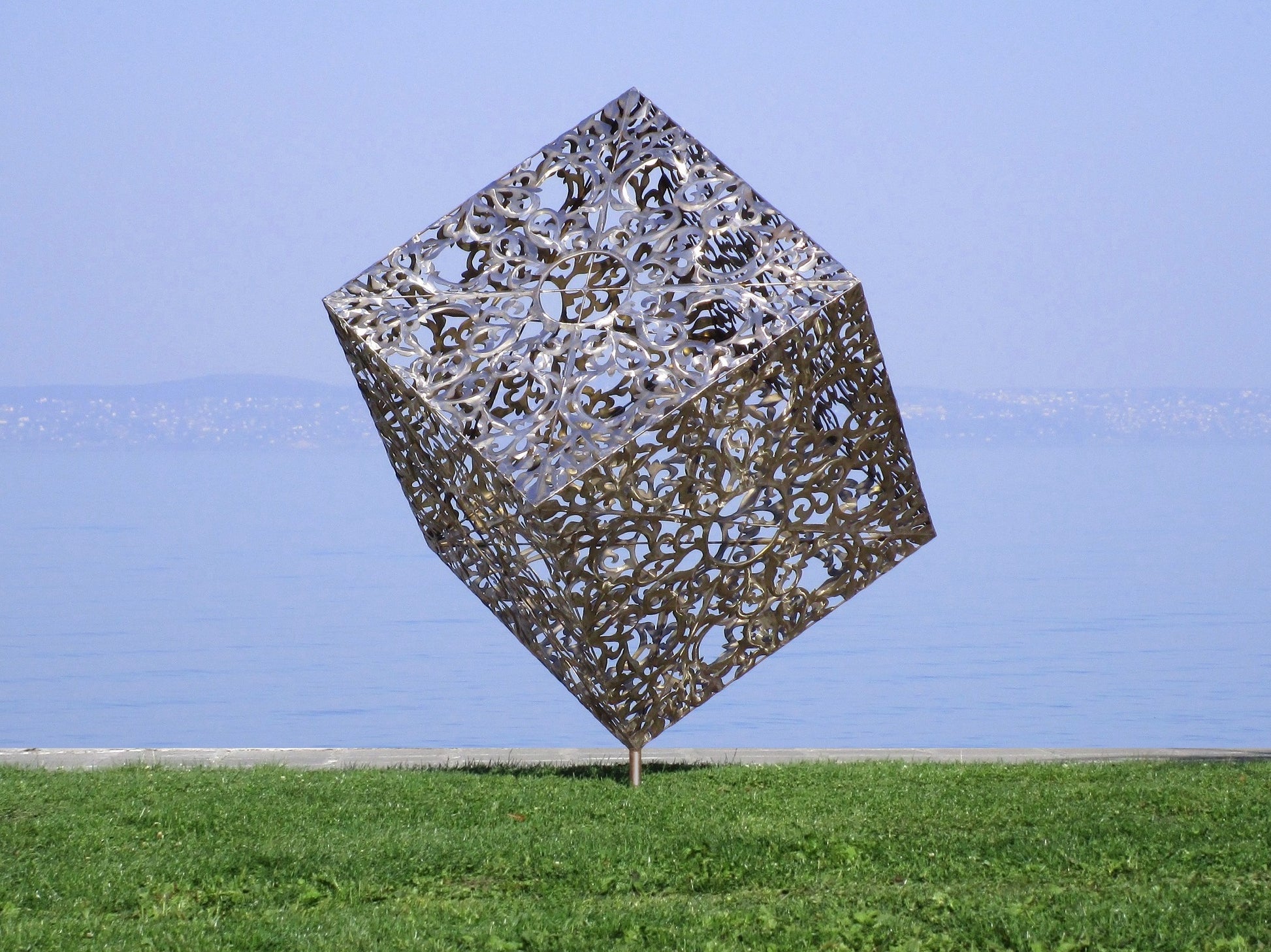 Moderne Gartenskulptur aus poliertem Edelstahl: Ein filigran durchbrochener Metallwürfel auf einer hohen, rostbraunen Cortenstahl-Stele direkt am Seeufer.