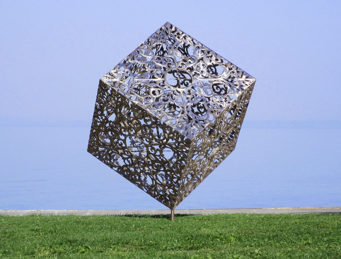 Detailansicht einer dekorativen Metallskulptur: Ein auf der Spitze stehender Würfel mit komplexen Ornamenten auf einer grünen Wiese vor Seekulisse.